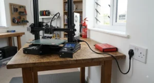 An FDM 3D printer plugged securely into a standard UK three-pin wall socket in a well-ventilated room